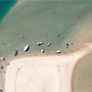 Vista aérea de uma estreita faixa de areia entre águas de rio e mar com barcos dispersos.